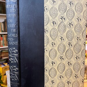 May include: A black hardback book with the title "Selected Tales of Guy de Maupassant" on the spine. The cover features a decorative pattern of grey leaves on a beige background.