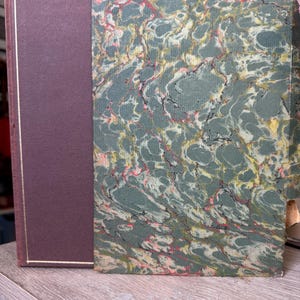 May include: A hardcover book featuring a solid dark maroon spine and cover section with a thin gold border line. The front cover panel displays a marbled paper design in shades of sage green, cream, and black with subtle accents of pink.