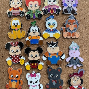 May include: A collection of sixteen enamel pins featuring stylized cartoon characters in a seated position. The pins include various figures with glittery accents, vibrant colors, and expressive faces, arranged in a grid on a cork board surface.