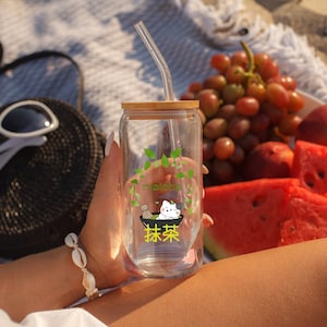 Puede incluir: Un vaso de vidrio transparente con tapa de bamb&uacute; y una pajita de pl&aacute;stico transparente. El vaso tiene un gr&aacute;fico verde y blanco de un gato bebiendo matcha. El texto "matcha" y "抹茶" est&aacute; impreso en el vaso.