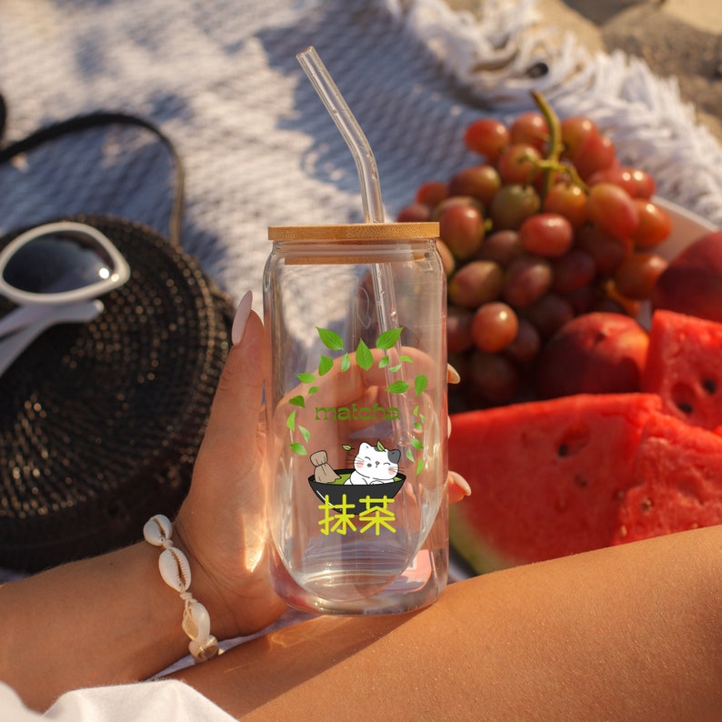 Puede incluir: Un vaso de vidrio transparente con tapa de bamb&uacute; y una pajita de pl&aacute;stico transparente. El vaso tiene un gr&aacute;fico verde y blanco de un gato bebiendo matcha. El texto "matcha" y "抹茶" est&aacute; impreso en el vaso.