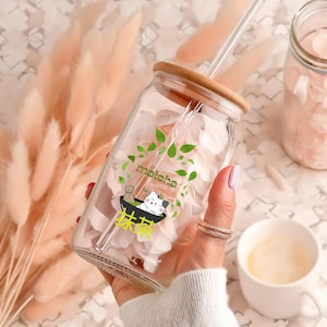 Puede incluir: Un tarro de vidrio transparente con tapa de bamb&uacute; y una pajita. El tarro est&aacute; lleno de p&eacute;talos de flores rosas y tiene una pegatina con un gato de dibujos animados y la palabra "matcha" en &eacute;l.