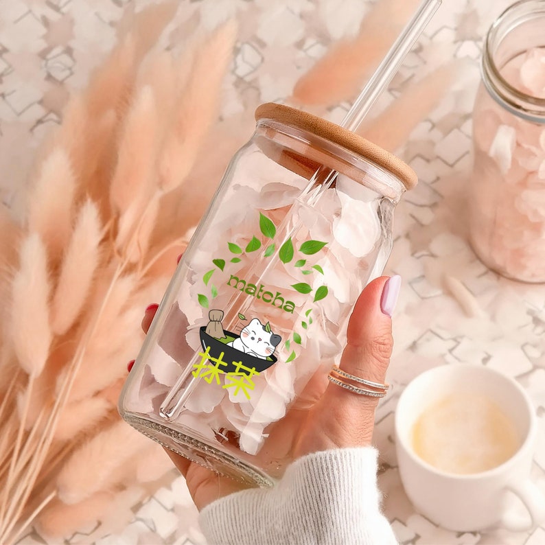 Puede incluir: Un tarro de vidrio transparente con tapa de bamb&uacute; y una pajita. El tarro est&aacute; lleno de p&eacute;talos de flores rosas y tiene una pegatina con un gato de dibujos animados y la palabra "matcha" en &eacute;l.