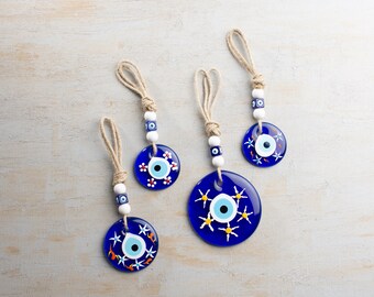 Op de afbeelding: Vier blauwe en witte boze oog charmes met witte kralen en touw dat eraan hangt. De charmes zijn versierd met witte en gele bloemen.
