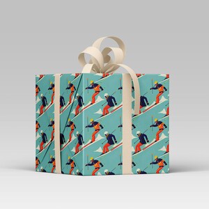 May include: A wrapped gift box featuring a repeating pattern of skiers on a light blue background. The skiers are depicted in a retro style, wearing orange and blue ski suits. The box is tied with a cream-colored ribbon and bow.