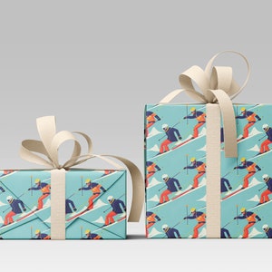 May include: Two gift boxes wrapped in light blue paper with a repeating pattern of skiers on a snowy mountain. The skiers wear orange and blue outfits. The boxes are tied with cream-colored ribbon.