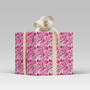 May include: A wrapped gift box with a cream-colored bow. The box is covered in a pink and red pattern of branches, ornaments, and flowers. Cream-colored ribbon wraps around the box. The gift box is on a white surface.