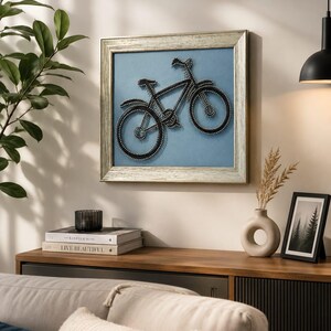 May include: A framed artwork featuring a black bicycle design made with string art on a light blue background. The frame is silver-toned, and the artwork is displayed on a wall above a wooden cabinet. Decorative items include books, a vase, and a framed picture.