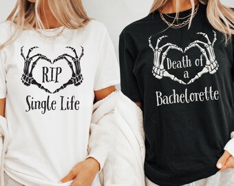 May include: Two white t-shirts with black skeleton hands forming a heart shape. One shirt says "RIP Single Life" and the other says "Death of a Bachelorette".