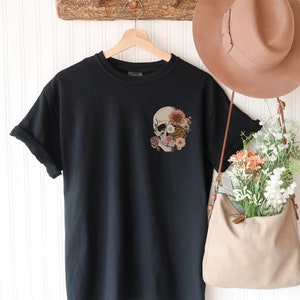 May include: Black t-shirt with a pocket featuring a skull with flowers covering the eye sockets.