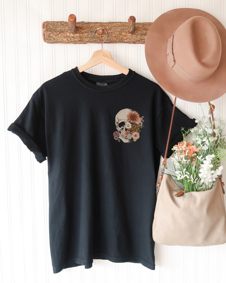 May include: Black t-shirt with a pocket featuring a skull with flowers covering the eye sockets.