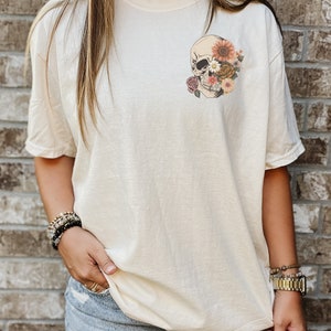 May include: A cream colored t-shirt with a floral skull graphic on the left chest. The skull is white with black eyes and is surrounded by pink, orange, and yellow flowers.