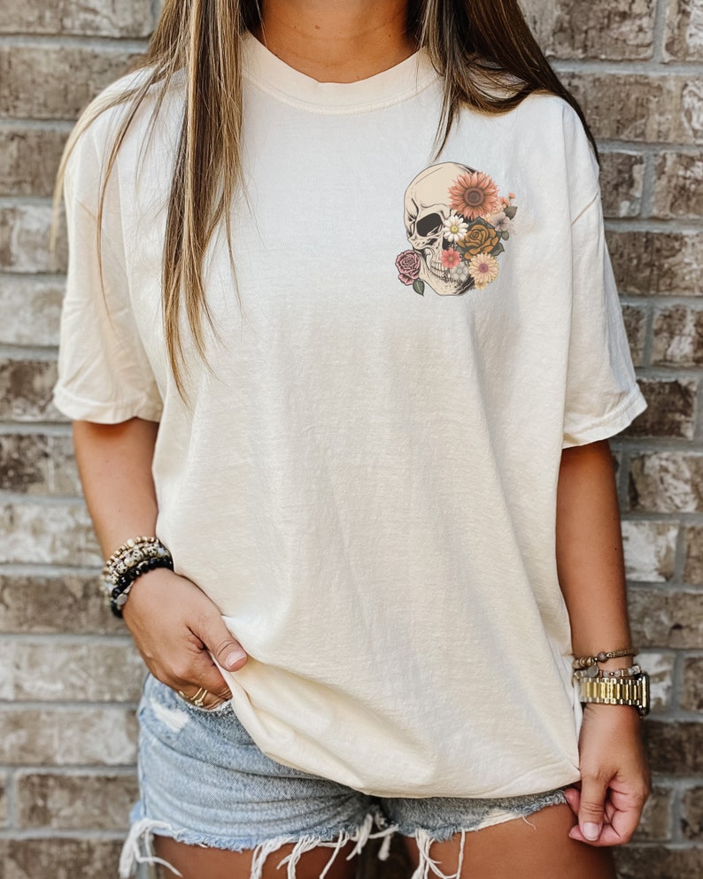 May include: A cream colored t-shirt with a floral skull graphic on the left chest. The skull is white with black eyes and is surrounded by pink, orange, and yellow flowers.
