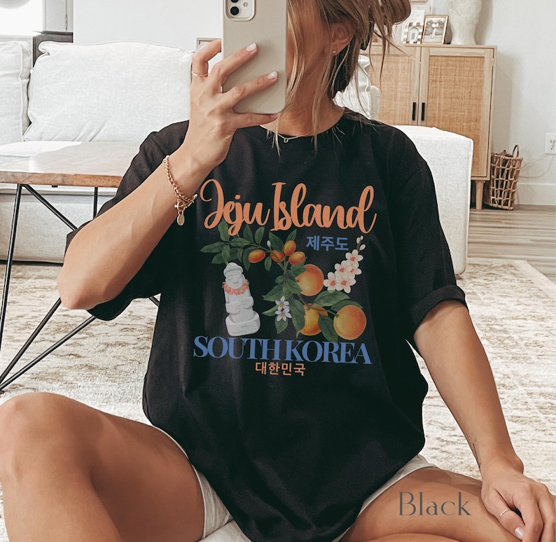 Puede incluir: Camiseta negra con un gr&aacute;fico de la isla de Jeju. El dise&ntilde;o presenta texto, ilustraciones de naranjas, flores y una estatua de piedra. Las palabras "Jeju Island" y "South Korea" est&aacute;n impresas en la camiseta.