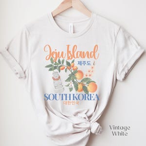 Puede incluir: Camiseta blanca vintage con un dise&ntilde;o de la isla de Jeju. El gr&aacute;fico incluye texto naranja, ilustraciones de naranjas, flores y una estatua de piedra, con las palabras "Jeju Island" y "South Korea" en ingl&eacute;s y coreano.