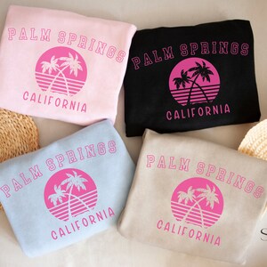 Puede incluir: Cuatro sudaderas de diferentes colores con el texto "Palm Springs California" y un gr&aacute;fico de palmera rosa.