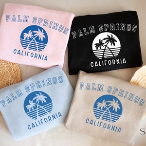 Puede incluir: Cuatro sudaderas de diferentes colores con un gr&aacute;fico de palmeras y el texto "PALM SPRINGS CALIFORNIA". Los colores son rosa, negro, azul claro y beige.