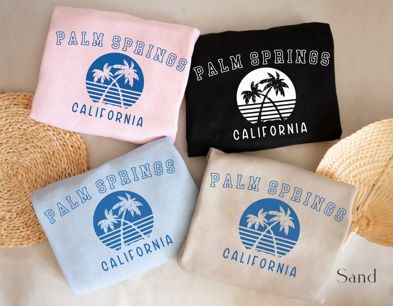Puede incluir: Cuatro sudaderas de diferentes colores con un gr&aacute;fico de palmeras y el texto "PALM SPRINGS CALIFORNIA". Los colores son rosa, negro, azul claro y beige.