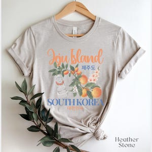 Puede incluir: Camiseta de color piedra de brezo con un dise&ntilde;o gr&aacute;fico. El dise&ntilde;o presenta las palabras "Jeju Island" en naranja, texto coreano y las palabras "South Korea". El dise&ntilde;o incluye ilustraciones de melocotones, flores y una estatua tradicional.