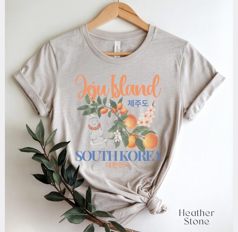 Puede incluir: Camiseta de color piedra de brezo con un dise&ntilde;o gr&aacute;fico. El dise&ntilde;o presenta las palabras "Jeju Island" en naranja, texto coreano y las palabras "South Korea". El dise&ntilde;o incluye ilustraciones de melocotones, flores y una estatua tradicional.