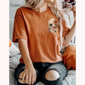 May include: A rust-colored t-shirt with a graphic of a skull with flowers covering one eye. The skull is in a vintage style with a floral design.