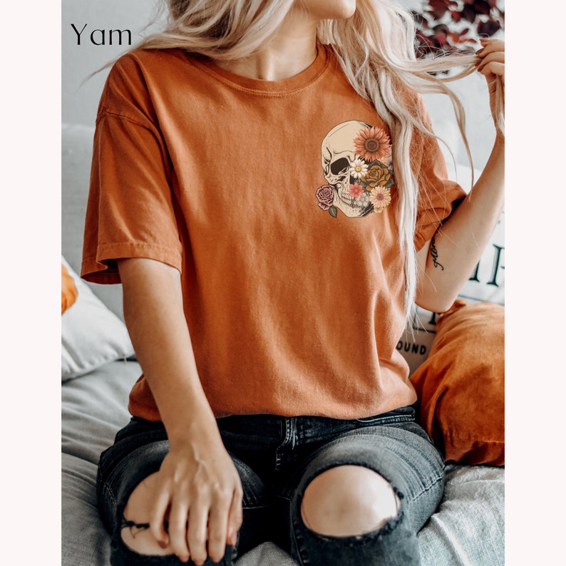 May include: A rust-colored t-shirt with a graphic of a skull with flowers covering one eye. The skull is in a vintage style with a floral design.