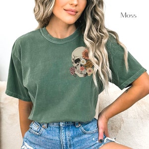 May include: A moss green t-shirt with a floral skull graphic. The skull is white with black eyes and a pink nose. The flowers are pink, yellow, and orange.