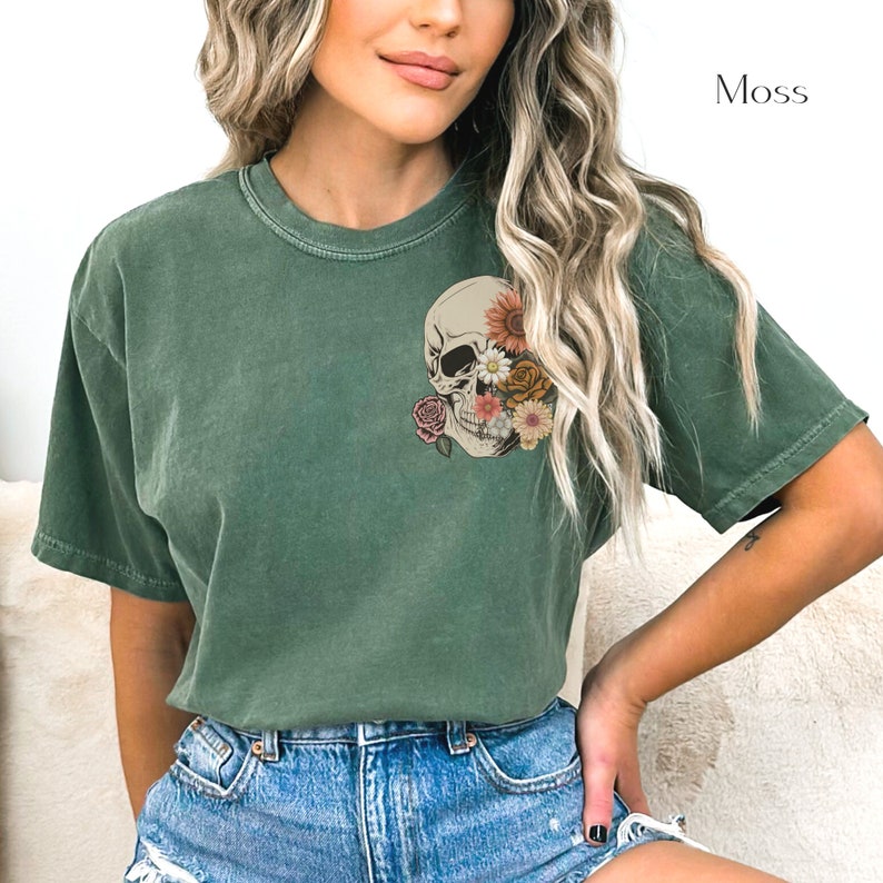 May include: A moss green t-shirt with a floral skull graphic. The skull is white with black eyes and a pink nose. The flowers are pink, yellow, and orange.