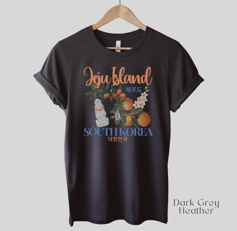 Puede incluir: Camiseta gris oscuro jaspeado con un gr&aacute;fico que presenta las palabras "Jeju Island" en escritura naranja, con texto coreano e ilustraciones de naranjas, flores y una estatua de piedra. Las palabras "South Korea" son azules.