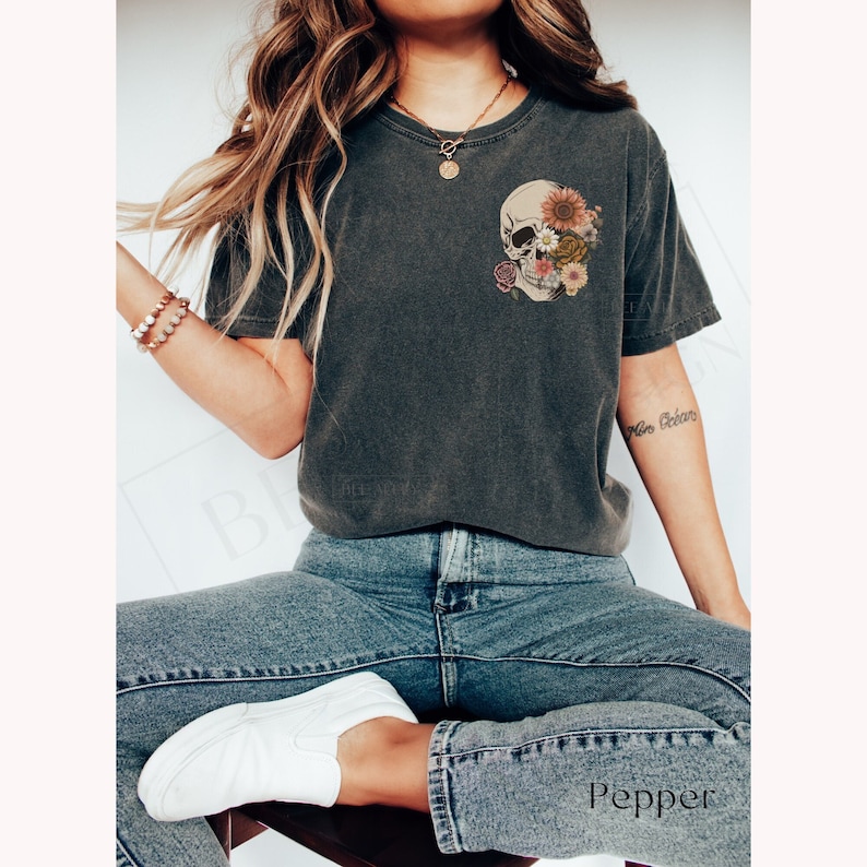 May include: A dark gray t-shirt with a floral skull graphic on the front. The skull is made of flowers and leaves in shades of pink, yellow, and green. The model is wearing the shirt with blue jeans and white sneakers.