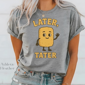 May include: Athletic heather gray t-shirt with the text "Later, Tater" and a cartoon hash brown character waving. The hash brown is yellow with a smiling face and brown details. The shirt is a casual style.