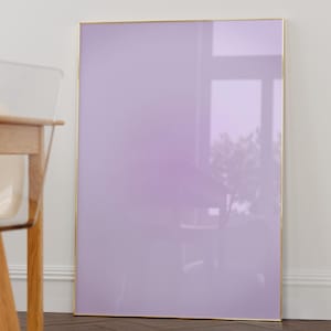 May include: A light purple framed picture with a gold frame is leaning against a white wall. The picture is in a room with a wooden floor and a wooden chair.