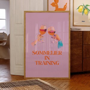 May include: A pink poster with two glasses of red wine clinking together. The text "SOMMELIER IN TRAINING" is printed in orange letters.