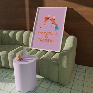May include: A pink framed print with a graphic of two wine glasses clinking. The text on the print reads "Sommelier in Training".
