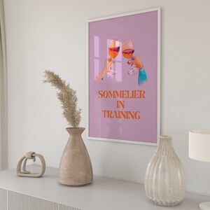 May include: A pink poster with a graphic of two hands holding glasses of wine. The text on the poster reads "Sommelier in Training".