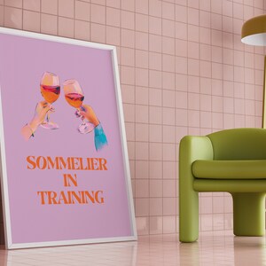 May include: A framed poster with a light purple background featuring two hands holding glasses of wine and the text "SOMMELIER IN TRAINING" in orange.