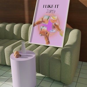 May include: A pink poster with the text "I like it dirty" in black lettering. Three hands are holding up glasses of cocktails. The poster is framed in white and is leaning against a green sofa.