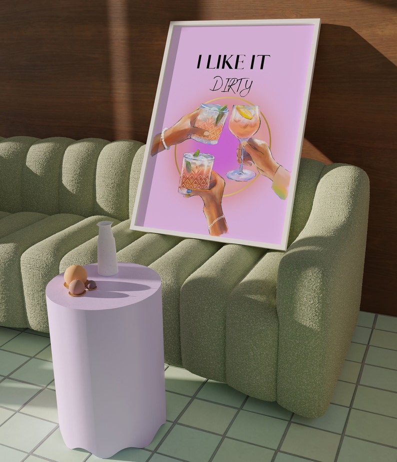 May include: A pink poster with the text "I like it dirty" in black lettering. Three hands are holding up glasses of cocktails. The poster is framed in white and is leaning against a green sofa.
