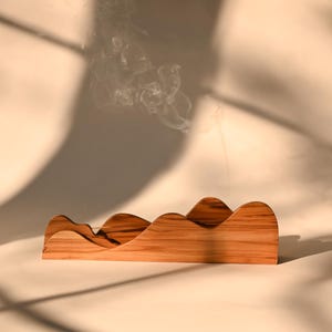 May include: A light brown wooden incense holder with a wavy, mountain-like silhouette. The piece features a natural wood grain pattern and a thin slot along the top edge. A single incense stick is inserted, emitting a thin trail of white smoke.