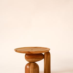 May include: A small wooden pedestal stand featuring a circular top surface, a central oval-shaped support, a rounded base, and a single vertical leg. The object is crafted from natural wood with a visible grain pattern and a smooth, polished finish.