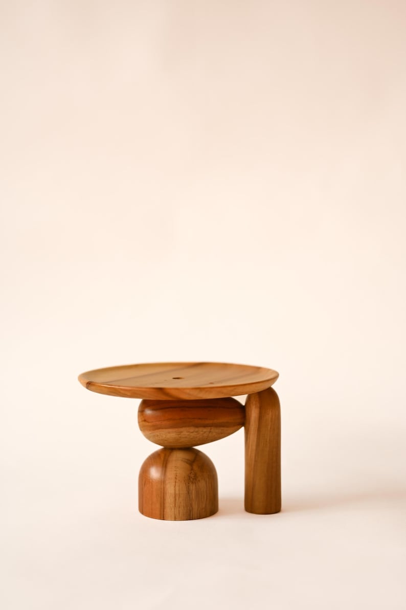 May include: A small wooden pedestal stand featuring a circular top surface, a central oval-shaped support, a rounded base, and a single vertical leg. The object is crafted from natural wood with a visible grain pattern and a smooth, polished finish.