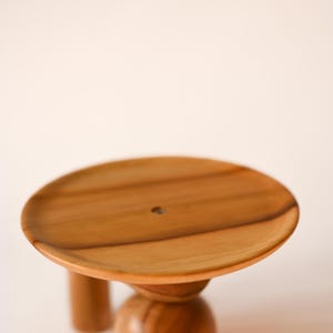 May include: A circular wooden stand featuring a smooth, light brown grain pattern on the flat top surface. The base consists of a central rounded pedestal and a secondary cylindrical support leg, both crafted from the same polished wood material.