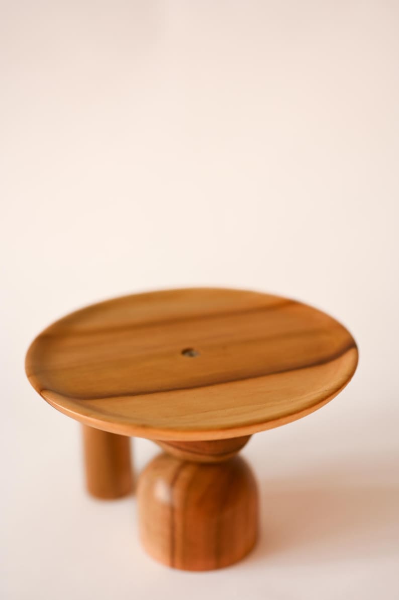 May include: A circular wooden stand featuring a smooth, light brown grain pattern on the flat top surface. The base consists of a central rounded pedestal and a secondary cylindrical support leg, both crafted from the same polished wood material.