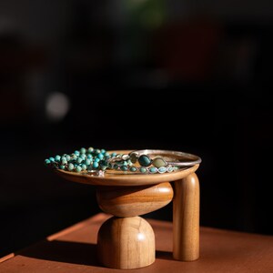 May include: A small wooden pedestal stand with a circular top and rounded base supports a collection of jewelry, including a strand of turquoise beads, a silver-toned metal bangle, and a thin ring with small blue stone accents.