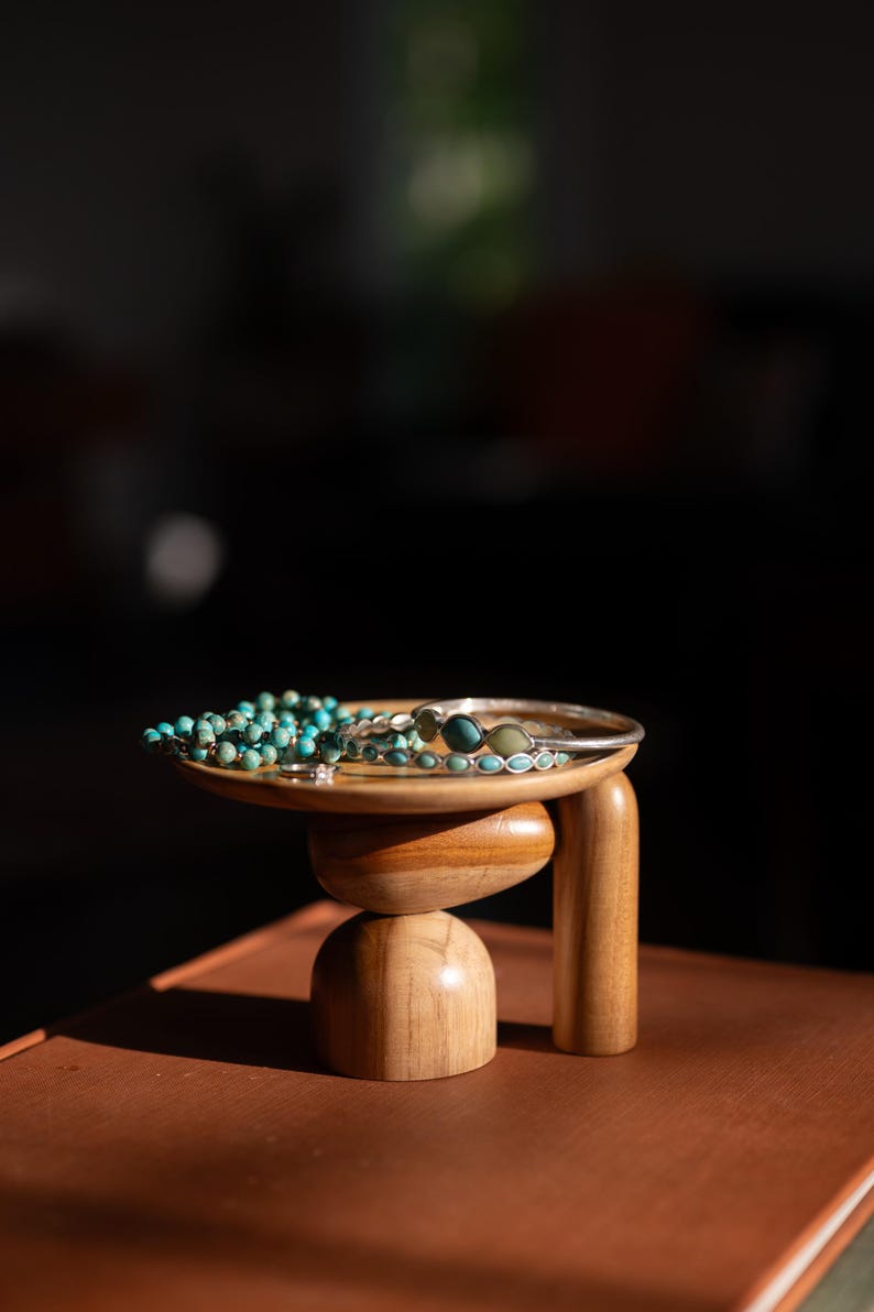 May include: A small wooden pedestal stand with a circular top and rounded base supports a collection of jewelry, including a strand of turquoise beads, a silver-toned metal bangle, and a thin ring with small blue stone accents.