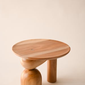 May include: A small wooden pedestal stand featuring a smooth, circular top surface with a visible metal screw in the center. The base consists of one cylindrical leg and one stacked support structure composed of a rounded, dome-shaped bottom piece.