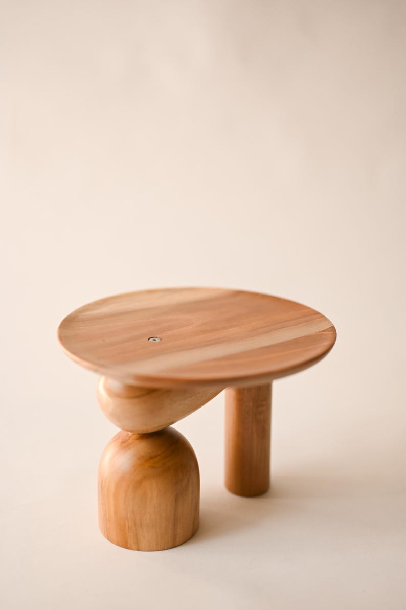 May include: A small wooden pedestal stand featuring a smooth, circular top surface with a visible metal screw in the center. The base consists of one cylindrical leg and one stacked support structure composed of a rounded, dome-shaped bottom piece.