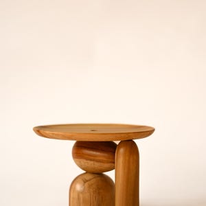 May include: A wooden display stand featuring a flat circular top supported by a central spherical element, a semi-spherical base, and a single vertical cylindrical leg. The object is crafted from light brown wood with a visible natural grain pattern.