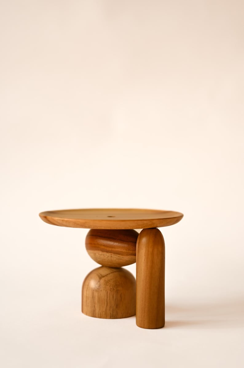May include: A wooden display stand featuring a flat circular top supported by a central spherical element, a semi-spherical base, and a single vertical cylindrical leg. The object is crafted from light brown wood with a visible natural grain pattern.