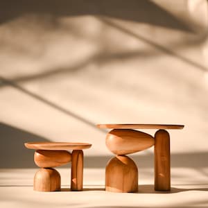 May include: Two wooden display stands of varying heights, each featuring a flat circular top supported by a base composed of a smooth, rounded dome-shaped piece, an oval-shaped element, and a single vertical cylindrical pillar.